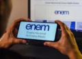 Mãos segurando smartphone com logo do Enem na tela azul clara, notebook com página oficial do exame ao fundo.