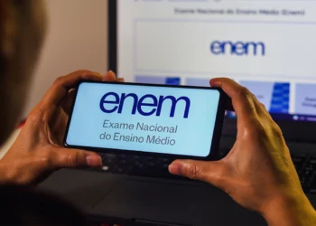 Mãos segurando smartphone com logo do Enem na tela azul clara, notebook com página oficial do exame ao fundo.