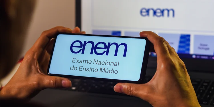 Mãos segurando smartphone com logo do Enem na tela azul clara, notebook com página oficial do exame ao fundo.
