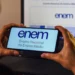 Mãos segurando smartphone com logo do Enem na tela azul clara, notebook com página oficial do exame ao fundo.