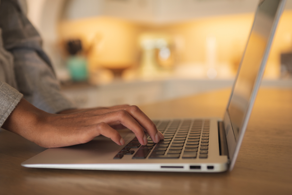 Inscrição Câmara Rio das Ostras RJ Mão feminina digitando em teclado de notebook prateado sobre mesa de madeira com fundo desfocado em tons quentes