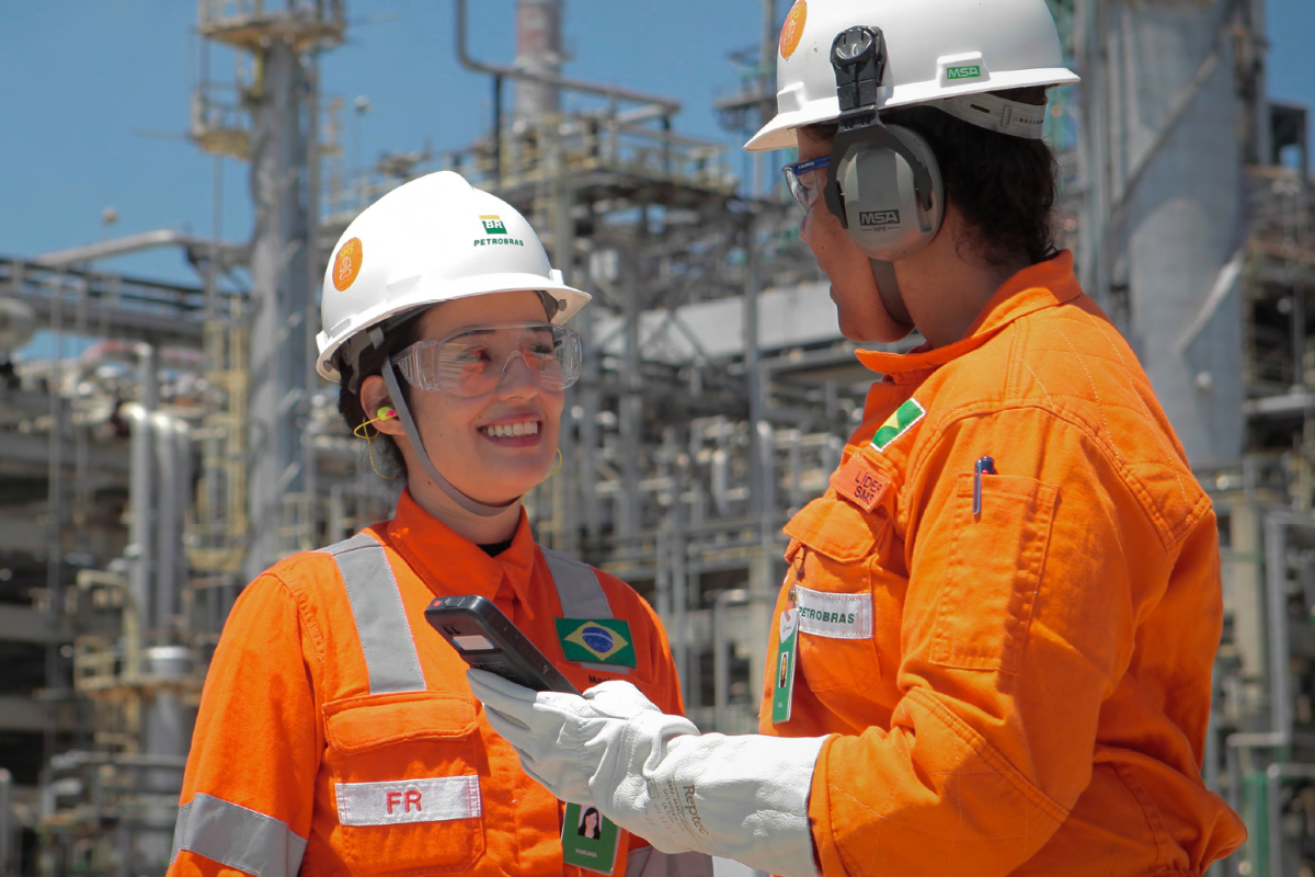 Inscrição Petrobras 2026 Duas profissionais da Petrobras em uniforme laranja e EPI conversam em refinaria, segurando equipamento de comunicação.