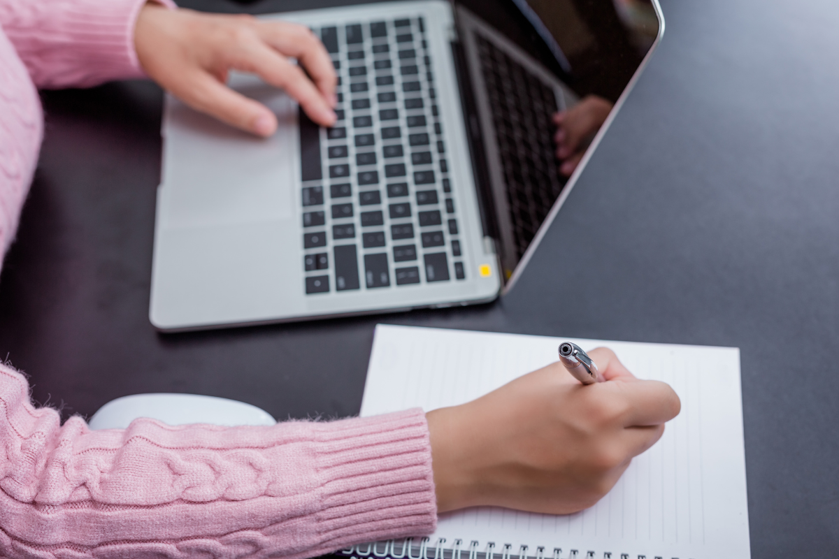 Inscrição Prefeitura Mãos com suéter rosa tricotado segurando caneta sobre caderno pautado ao lado de laptop prateado em mesa escura.