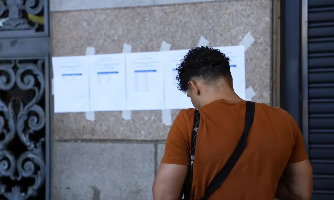 Local de atuação CNU 2025 Jovem de camiseta laranja e mochila preta confere lista de aprovados afixada em parede de pedra com fita